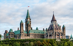 Wide view of the Canadian Parliament buildings in Ottawa, with a green copper roof, pointed towers, and a large central clock tower. The building is set against a partly cloudy sky and surrounded by green foliage in the foreground.